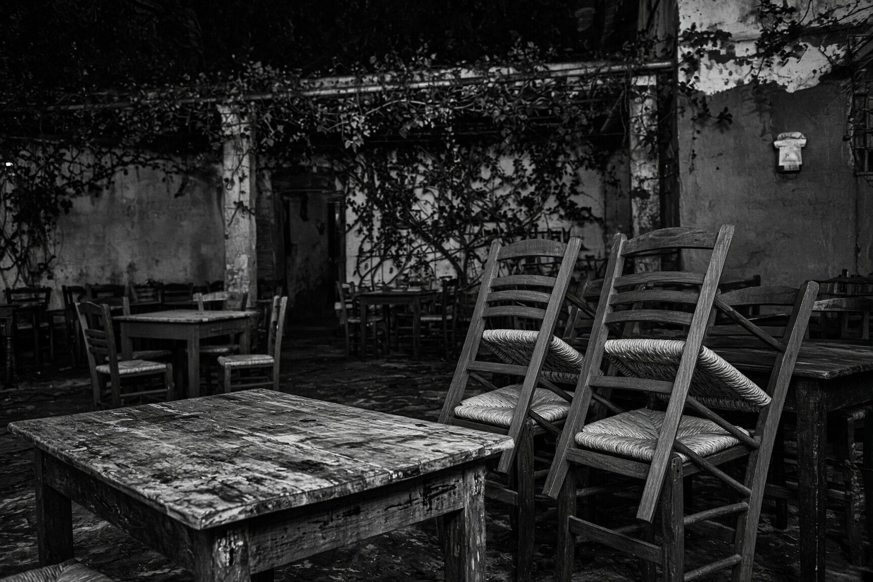 The courtyard before service — a scarred table in the foreground, rush-seat chairs tipped against it, the vine overhead — illustrative AI-generated demo image.